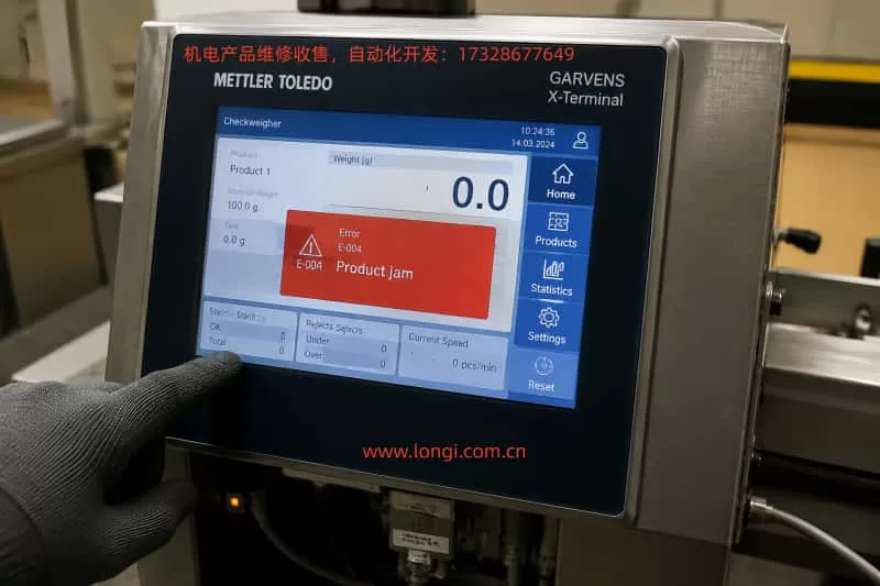 Close-up of a Mettler Toledo Garvens X-Terminal industrial touchscreen panel in a factory environment, displaying a red error message 'E-004 Product jam', with a gloved hand pointing at the screen and cables visible around the stainless steel enclosure.
